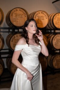 Bride sipping whiskey with Hush & Whisper Barrels behind her. 