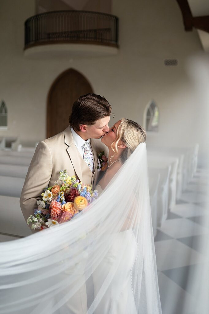 Bride and Groom kissing inside a chapel with a colorful bridal bouquet.